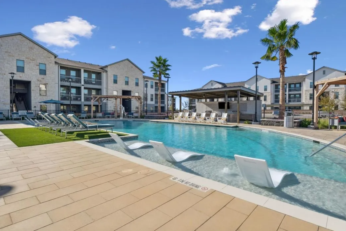 A photo of an apartment complex pool with luxurious lounge chairs set in front of a bright blue sky and palm trees.